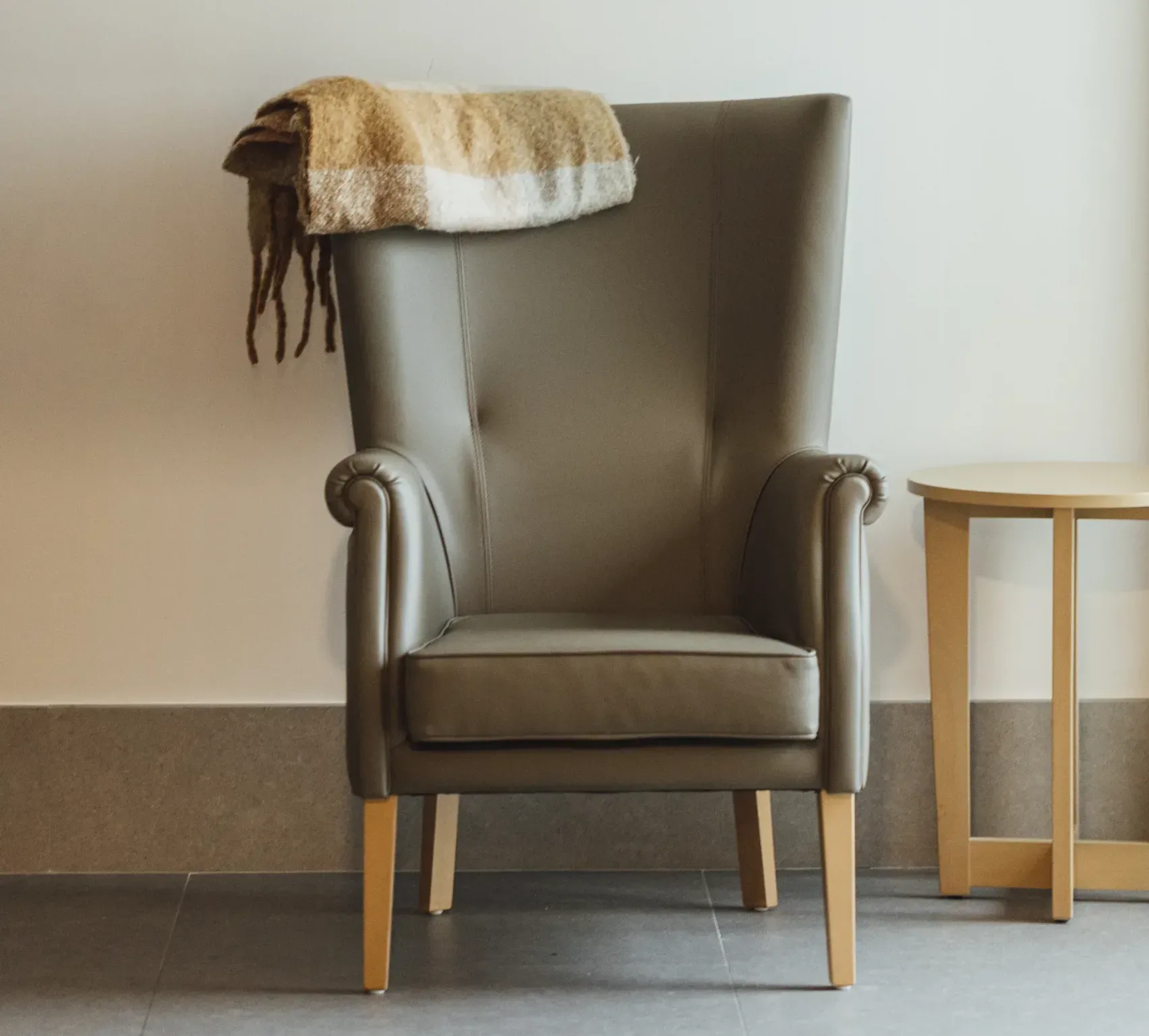 Butaca color beige de una habitación de residencia con una mantita y una mesita a su derecha